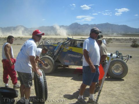 1990 Ford Bronco - Crew members stand with a spare tire beside a blue and yellow tube-frame race car in a dusty desert pit area.