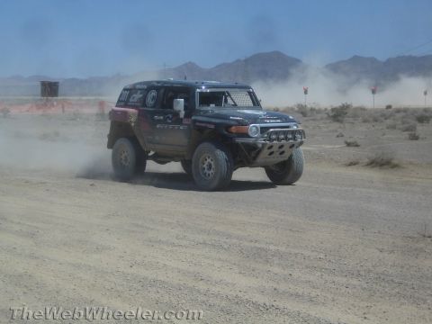 1990 Ford Bronco - black Toyota FJ Cruiser race vehicle kicks up dust as it races along a desert course with mountains behind.