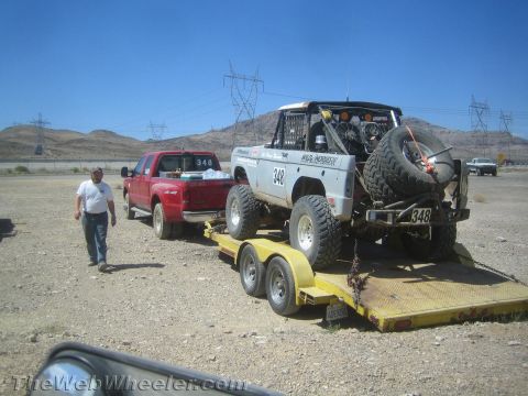 1990 Ford Bronco - man stands by a red pickup towing a white tube-chassis off-road race truck marked 348 on a flatbed trailer in the desert.
