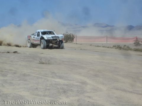 1990 Ford Bronco - white off-road race truck throws up a cloud of dust as it speeds along a desert course near a red fence.