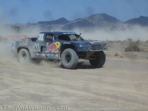 1990 Ford Bronco - blue Red Bull-liveried off-road race truck slides through a dusty desert corner.