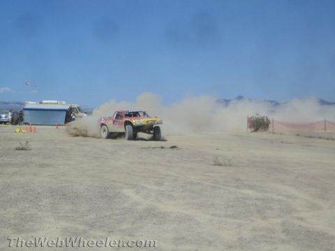 1990 Ford Bronco - off-road race truck slides through a dusty desert section past a parked trailer and traffic cones.