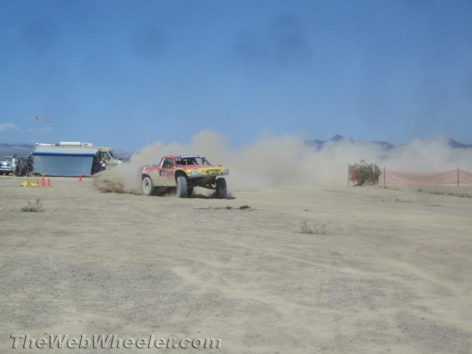 Off-road race truck sliding through dust past a trailer in the desert