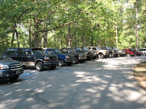 1995 Ford C-350 (Centurion Conversions) - A row of various Ford trucks and SUVs are parked in a lot surrounded by trees.