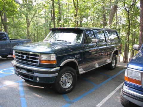 1995 Ford C-350 (Centurion Conversions) - A dark green Ford Bronco is parked in a handicapped spot, with a blue pickup truck partially visible to its right.