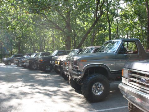 1995 Ford C-350 (Centurion Conversions) - A row of modified Ford trucks and Broncos are parked in a shaded, wooded area.