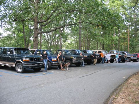 1995 Ford C-350 (Centurion Conversions) - A group of people stand with their Ford trucks and SUVs in a parking lot shaded by large trees.