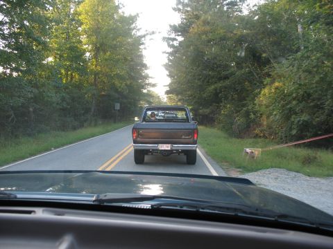 1995 Ford C-350 (Centurion Conversions) - A dark pickup truck is seen from behind on a rural road, with trees lining both sides.