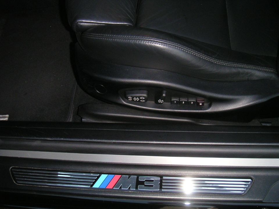 Close-up of a 2002 BMW M3 interior showing the door sill with M3 badging and part of the leather seat with memory controls