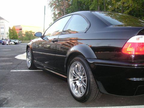 2002 BMW M3 - Black BMW coupe from the rear quarter, parked in a lot with buildings and trees in the background.