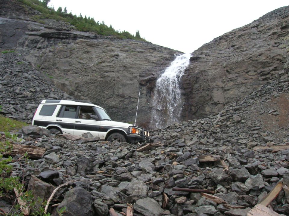 White Land Rover Discovery SUV parked on rocky slope below a waterfall