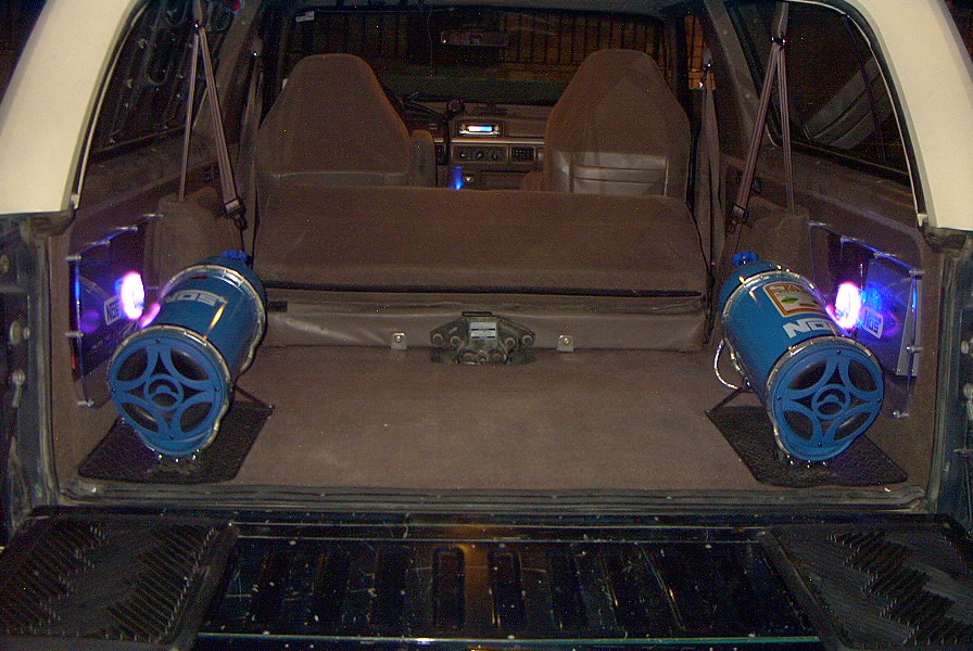 Vehicle interior showing two blue cylindrical speakers with purple lights, facing rearward. Seats and dashboard visible