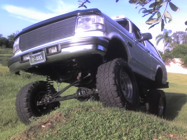 Silver lifted Ford Bronco with large tires on grassy hill, front grille and license plate visible