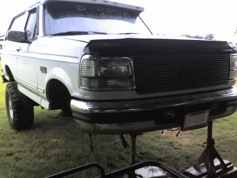1995 Ford Bronco - A white Ford Bronco is elevated on jack stands, showing its front end and large tires.