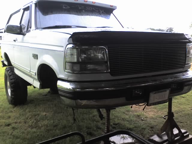White Ford Bronco on jack stands, front end visible. Hood is up, grille is blacked out. Large tires are mounted