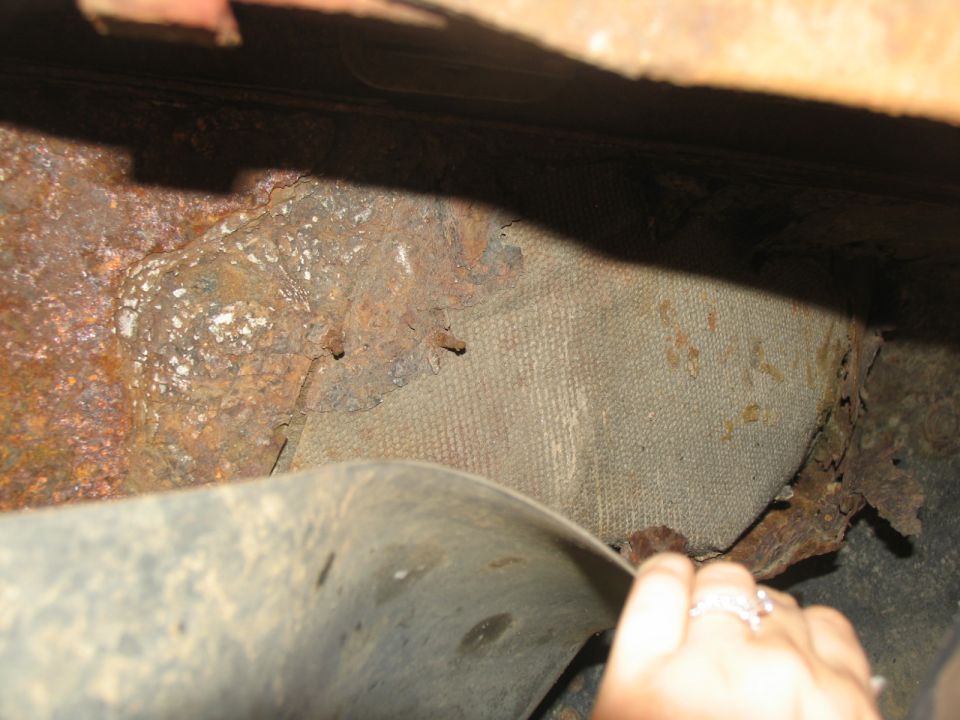 Close-up of heavily rusted metal and fabric material on a vehicle's undercarriage. A hand is visible near a curved metal part
