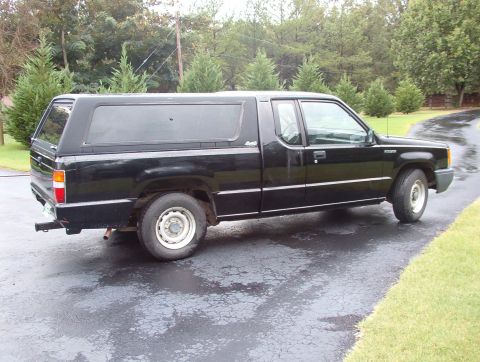 1993 Mitsubishi FTO - A black pickup truck with a camper shell is parked on a wet asphalt driveway, surrounded by trees.
