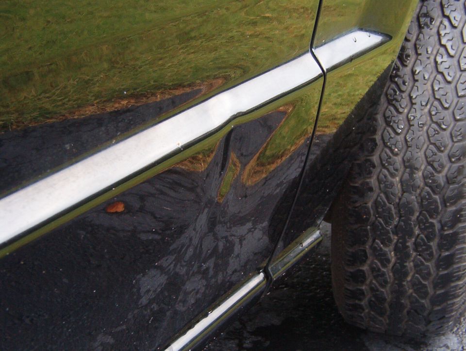 Black car's wet lower door panel with chrome trim, a dent, and a tire visible at right.