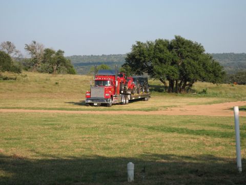2001 Jeep Wrangler - A red semi-truck transports two vehicles on a flatbed trailer across a grassy landscape under a clear blue sky.