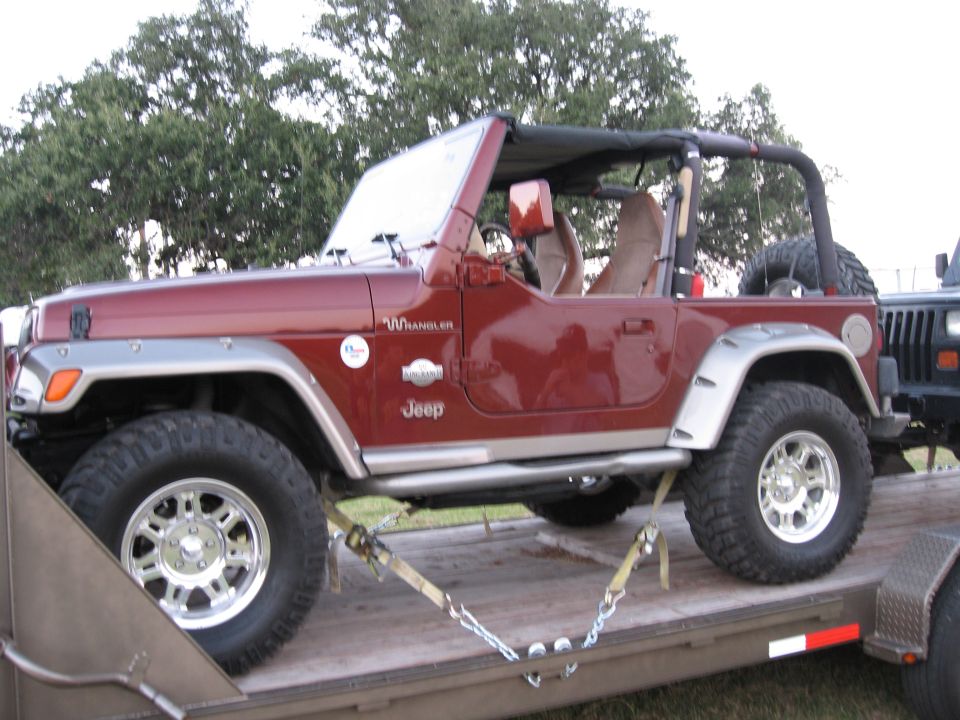 Red Jeep Wrangler on a trailer, showing the side and front quarter. It has a soft top and silver fender flares