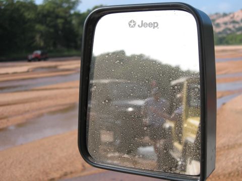2001 Jeep Wrangler - Reflection in a dirty Jeep side mirror shows a person and another vehicle in the background on a dirt road.