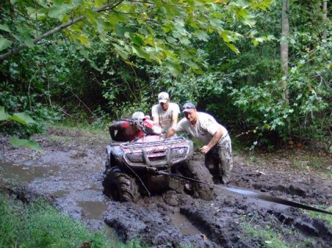 1992 Ford F-150 - Three people attempt to free a red ATV that is deeply stuck in thick mud on a wooded trail.
