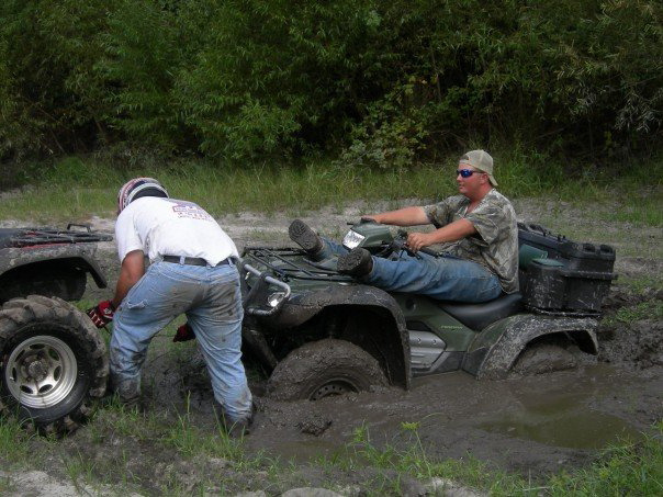 Two ATVs stuck in mud, one with a person sitting on it and another person trying to free it