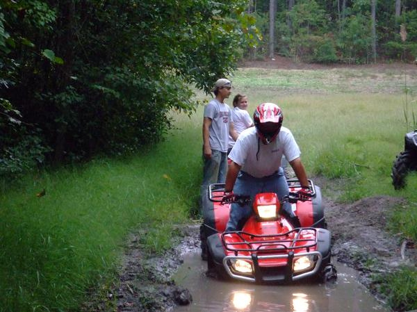 Red ATV stuck in mud with people watching. Another ATV is partially visible on the right