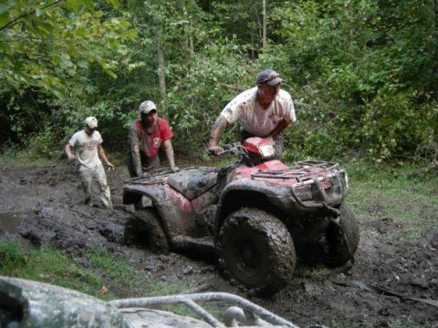 1992 Ford F-150 - Three people struggle to free a red ATV stuck deep in mud on a wooded trail.