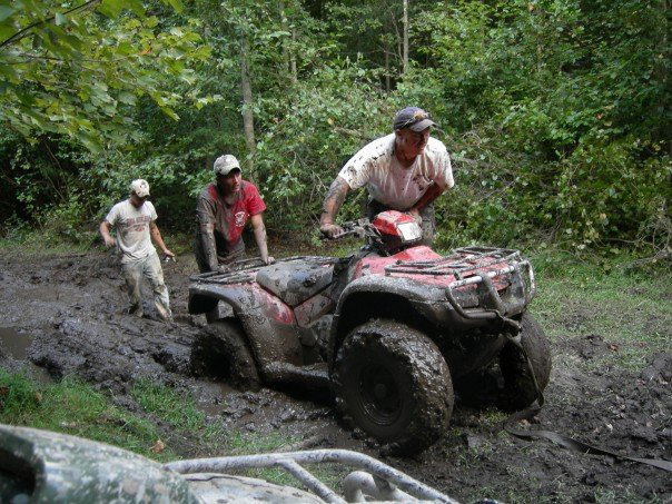 Red ATV stuck in mud, two people pushing it while another person sits on it. Another ATV is partially visible in the foreground