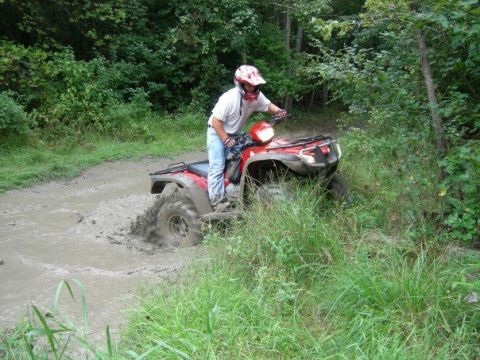 1992 Ford F-150 - A rider in a white T-shirt, jeans and a red-and-white helmet stands on the pegs of a red utility ATV as it climbs out of a muddy trail hole…