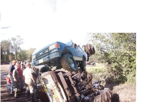 1987 Suzuki Samurai - A blue pickup truck is precariously balanced on another vehicle, stuck deep in the mud, while onlookers observe.
