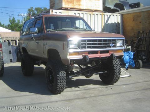 1990 Ford Bronco - brown two-tone lifted Ford Bronco II with mud tires and headlights on sits in a shop yard.