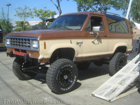 1990 Ford Bronco - brown-and-tan two-tone lifted Ford Bronco II on black wheels and BFGoodrich tires sits in a paved lot.