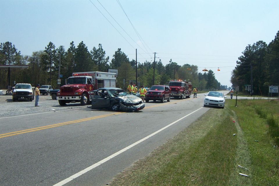 Car accident with damaged sedan, ambulance, and fire trucks on the side of the road. Emergency personnel are present