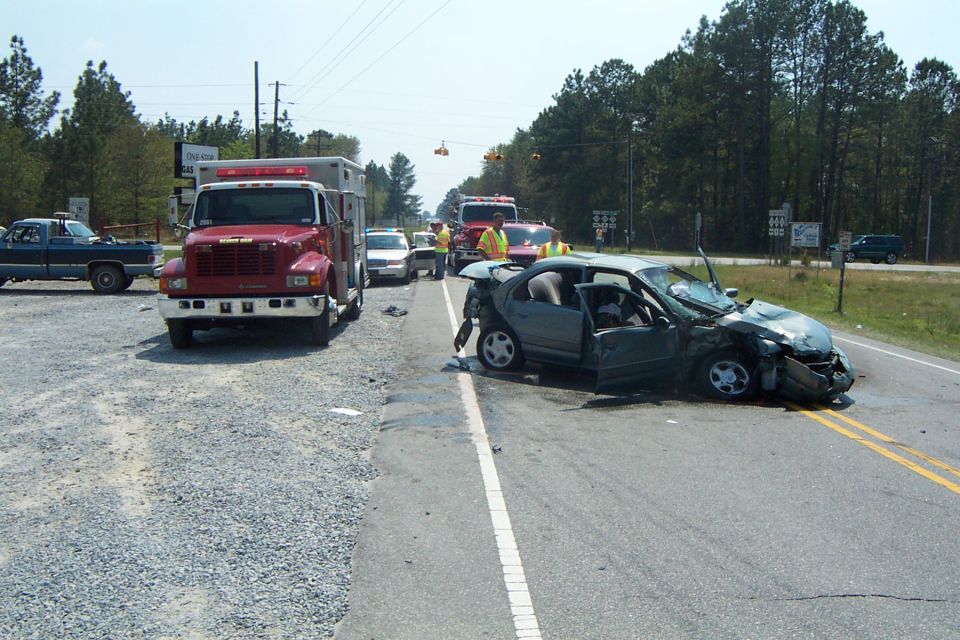 Damaged gray sedan with doors open, front end crushed, on a road with emergency vehicles