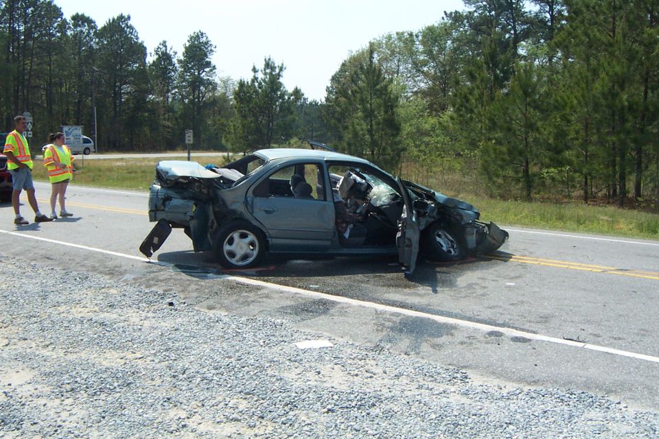 wrecked sedan on side of road, front end smashed, doors open, two people in safety vests standing nearby