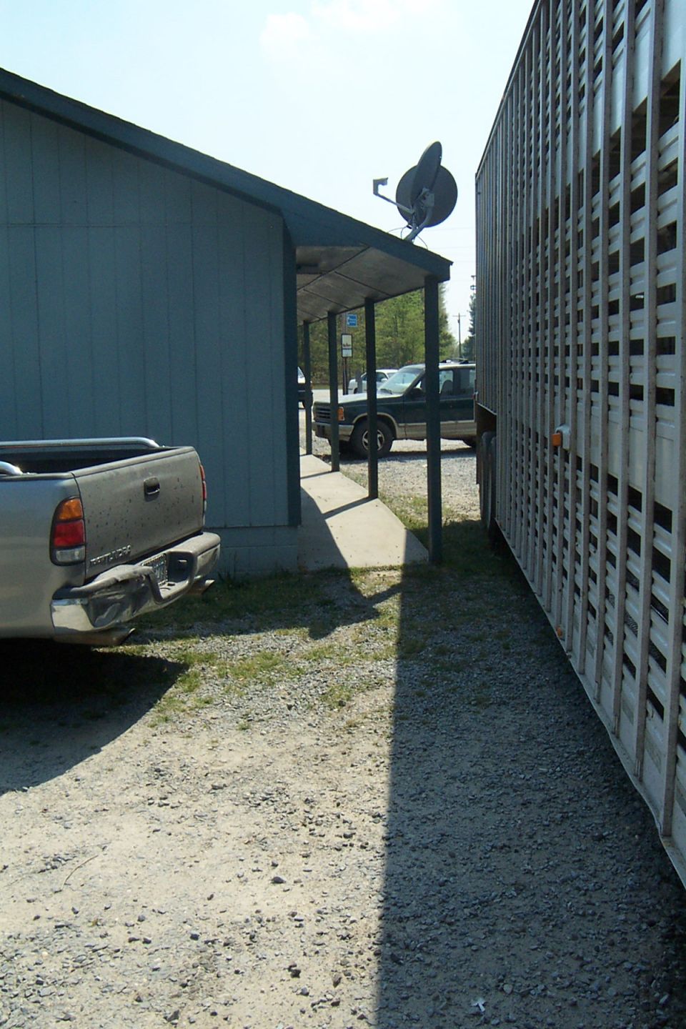 Silver pickup truck parked next to a large livestock trailer, with a building and another vehicle in the background