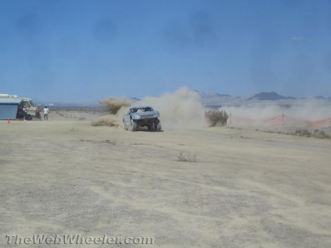 1990 Ford Bronco - off-road race truck launches a wall of dust as it powers across a desert course near a trailer.