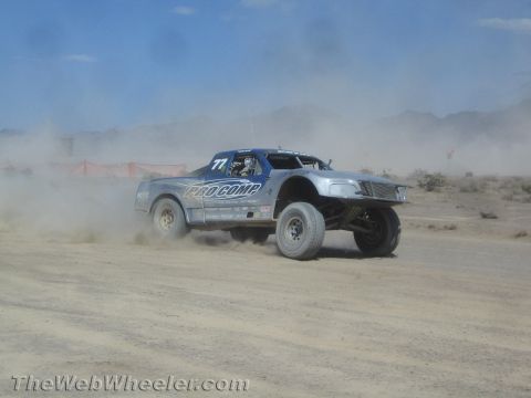 1990 Ford Bronco - silver off-road race truck numbered 77 with Pro Comp graphics speeds through a dusty desert course.