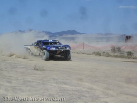 1990 Ford Bronco - blue off-road race truck slides through a dusty desert corner alongside a red course fence.