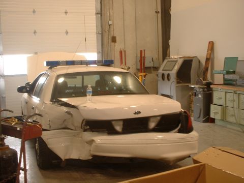 2000 Ford Crown Victoria - A damaged 2000 Ford Crown Victoria police car sits in a garage with its hood up and rear end crushed.