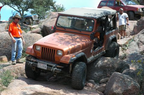 2001 Jeep Wrangler - An orange Jeep Wrangler tackles a challenging rocky trail, with other vehicles and people present.