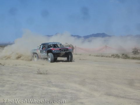1990 Ford Bronco - yellow-and-gray off-road race truck corners hard, kicking up heavy dust on a desert course.