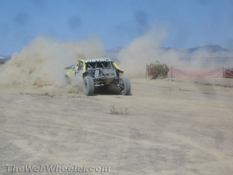1990 Ford Bronco - yellow off-road race truck slides through dust along a desert course marked by a red fence.