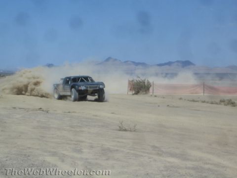 1990 Ford Bronco - off-road race truck slides through a cloud of dust on a desert course bordered by a red fence.