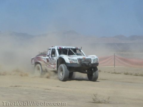 1990 Ford Bronco - white-and-red off-road race truck speeds through a dusty desert course near a red fence.