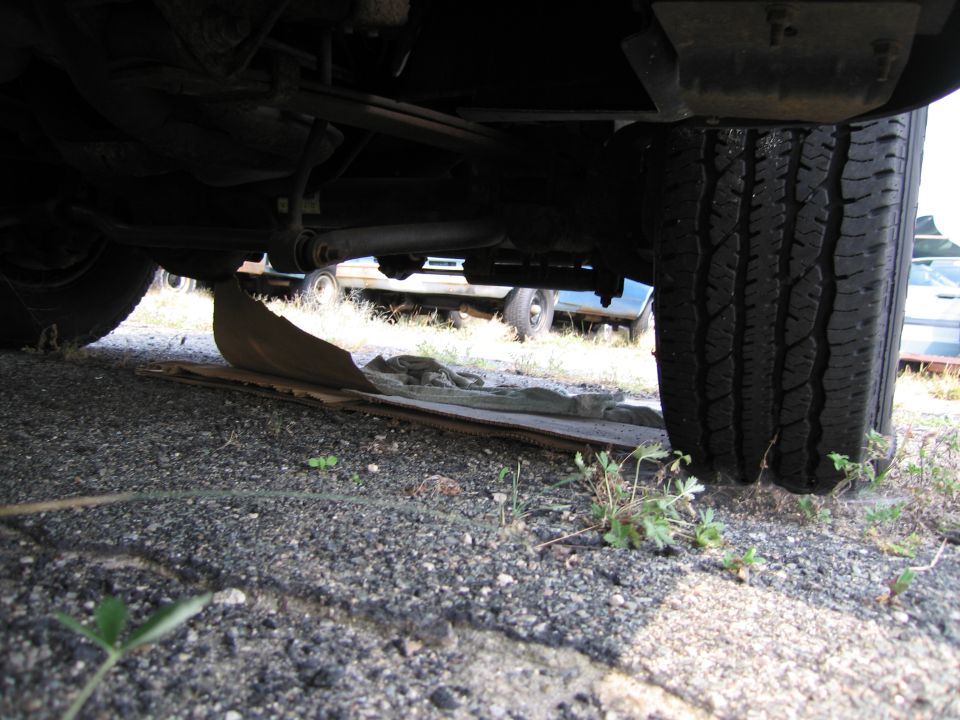 Underside view of a vehicle's rear axle, leaf spring, and tire. Other vehicles are visible in the background