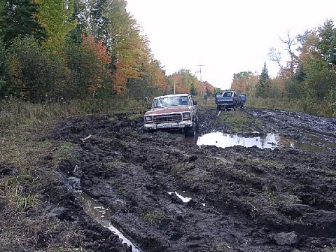 1978 Ford Bronco - A red 1978 Ford Bronco is stuck in deep mud on a trail, with another truck visible in the background.