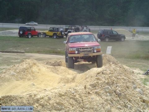 1995 Ford Bronco - Several off-road vehicles are gathered at an outdoor event, with one red SUV navigating a dirt obstacle.